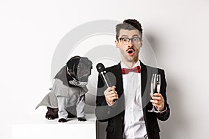 Male entertainer raising glass of champagne, giving microphone to cute black dog, standing over white background