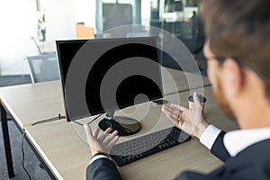 Male entrepreneur gesturing at computer webcamera, monitor with blank screen, man having meeting at workplace in office