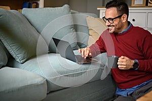 Male entrepreneur holding coffee cup and working over laptop on sofa while sitting in living room