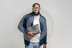 Male entrepreneur stands in studio isolated on gray and carrying trendy laptop