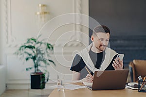 Male entrepreneur studying online, using smartphone while working at his workplace in modern office
