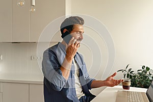 Male entrepreneur talking by phone while working on laptop at home kitchen. Distance work concept