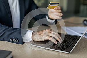 Male entrepreneur using credit card and laptop while sitting at workplace in office, focus on hand on keyboard