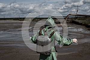 A male environmentalist in a green protective suit and gas mask takes a sample of water in a polluted lake. Waste from