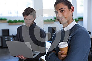 Male executive standing with arms crossed while his coworker using laptop