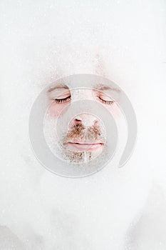 Male face in a lush foam, only eyes and mouth are visible, close-up. Man relaxes while lying in a bathtub, with soapy