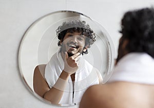 Male facial skincare concept. Young indian man touching beard, caring for skin in morning, standing in bathroom