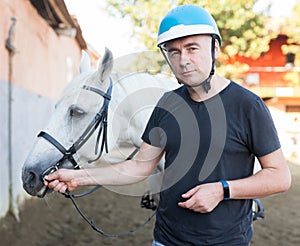 Male farmer in helmet standing with white horse at stable outdoor