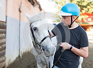 Male farmer in helmet standing with white horse at stable outdoor