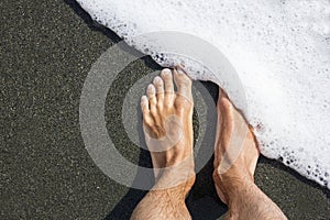 Male feet on the black volcanic sand covers with white sea foam. Minimalism. Geometric. Diagonal