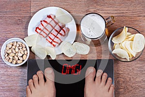 Male feet on digital scales with word omg on screen. Beer and plates with junk food sausages, potato chips, pistachios.