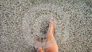 Male feet walking over sandy ocean beach and pebbles through calm clear sea water