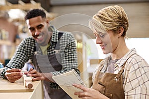 Male And Female Apprentices Working As Carpenters In Furniture Workshop Measure Wood And Take Notes