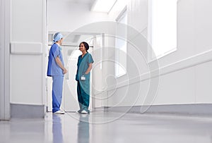 Male And Female Doctors Wearing Scrubs Meeting In Busy Hospital Corridor