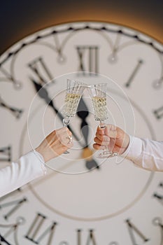 Male and female hands clink glasses of champagne in a living room decorated for Christmas. Big white clock, glasses of champagne.