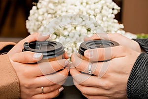 Male and female hands hold two brown glasses of coffee, next to a bouquet of gypsophila flowers