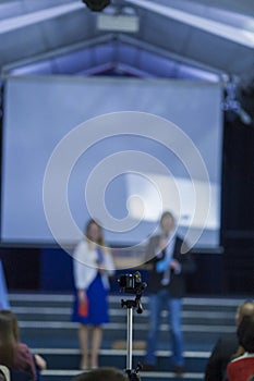 Male and Female Hosts on Stage In front of Group of People.