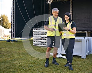 Male And Female Production Team With Headsets Setting Up Outdoor Stage For Music Festival Or Concert