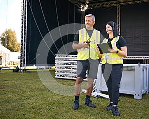 Male And Female Production Team With Headsets Setting Up Outdoor Stage For Music Festival Or Concert