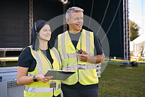 Male And Female Production Team With Headsets Setting Up Outdoor Stage For Music Festival Or Concert