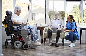 Male And Female Residents Sitting In Chair And Talking With Nurse In Retirement Home