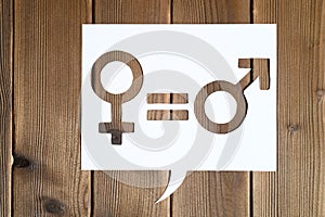 Male and female symbols and an equal sign in white dialogue box cut out of paper on a wooden background. the concept of gender equ