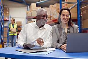 Male And Female Team Leaders Working On Laptop In Warehouse With Workers In Background