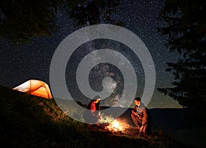 Male and female warming themselves around the fire at night camping above starry sky