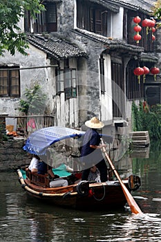 Male figure is seen in a small wooden rowboat driving people in it in a typical Asian evironment