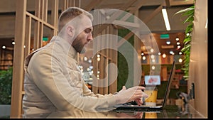 Male freelancer with beard sitting in cozy coworking cafe and working online with laptop, online business, programming