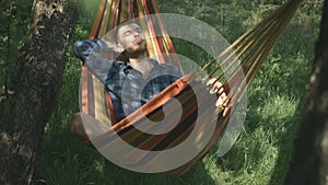 Male freelancer in casual wear lying in hammock at nature. Man with closed eyes relaxing on hammock outdoors. Male tourist enjoyin