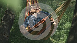 Male freelancer lying in hammock, relaxing and listening to music in earphones. Young relaxed man in earphones listens to music in