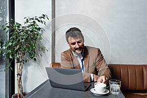 Male freelancer sitting at a restaurant table, drinking coffee and working on a laptop computer. Expatriate man in cafeteria doing