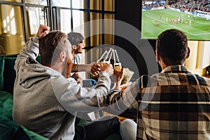 Male friends drinking beer and cheering with bottles while watching football match on TV
