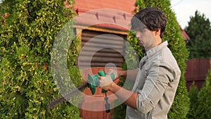 Male gardener is trimming an overgrown green thuja bush with an electric hedge clipper.