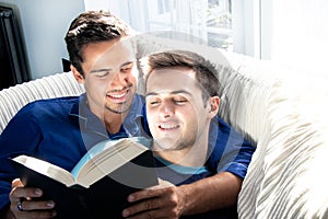 Male gay couple cuddling and reading together on large couch next to patio doors with bright sunlight shining on them