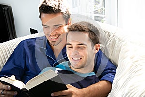 Male gay couple cuddling and reading together on large couch next to patio doors with bright sunlight shining on them