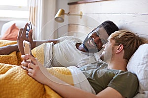 Male Gay Couple Lying In Bed At Home Checking Mobile Phones Together