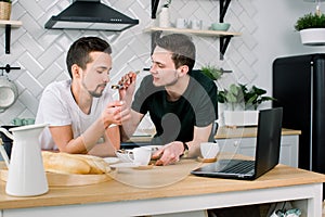 Male gay couple sitting at the table and having breakfast in their kitchen. Handsome young man in black t-shirt feeding