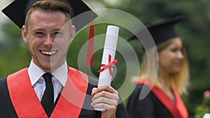 Male graduate in academic dress holding university diploma, smiling to camera