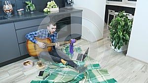 Male guitarist watching online guitar lessons and trying to play new chords. Man sitting on floor in kitchen and learning to play