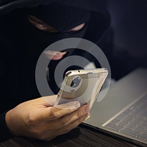 Male hacker in a black mask uses smartphone and laptop. A fraudster commits cyber crime. Fraudulent scheme with personal data and