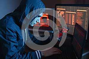 Male hacker in the hood holding the phone in hands trying to hack the mobile device cloud in the dark under neon light