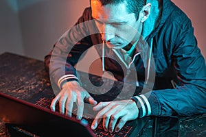 A male hacker is typing on a laptop keyboard in a dark room under a neon light. Cybercrime fraud and identity theft