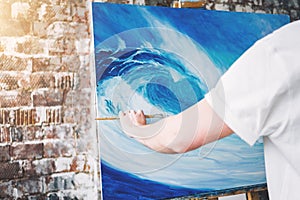 Male hand of artist holding paintbrush on background of canvas on easel. Professional painter drawing ocean wave in loft studio