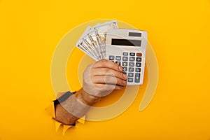 Male hand with calculator and dollar banknotes through a paper hole in yellow background