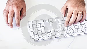 Male hand clicking on a mouse and typing on a white keyboard on a white table