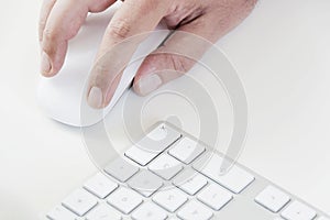 Male hand clicking on a white mouse with a white keyboard on the right. White table in a office