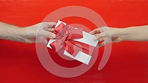 Male hand is giving present gift box to female hand. White gift box with red ribbon bow for Valentine`s Day, Christmas or Birthday