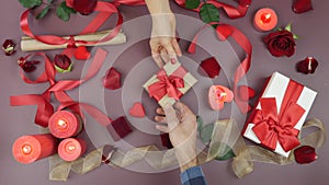 Male hand is giving wrapped gift box with tied red bow to female hand for St. Valentine`s Day against festive background with rose